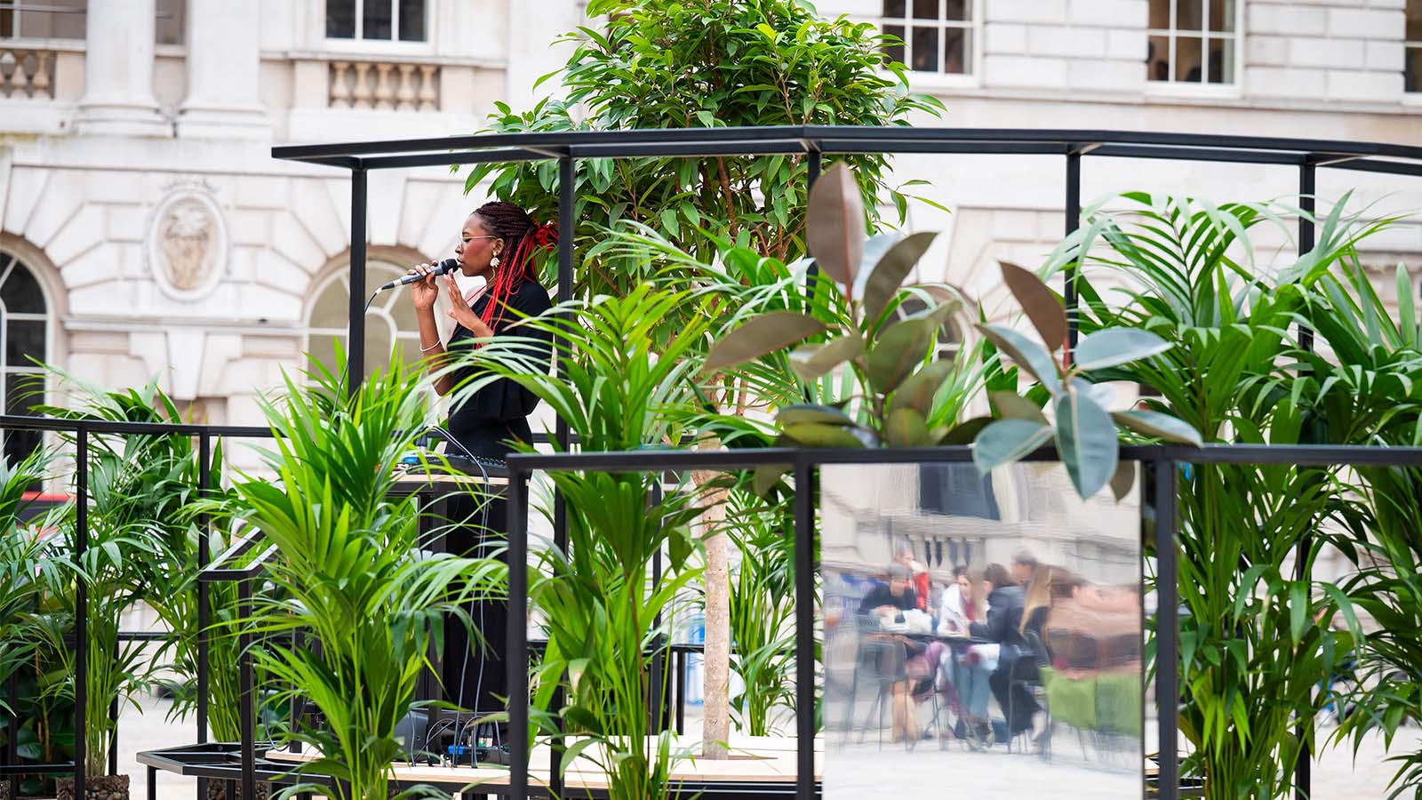 Woman singing into a microphone, standing on a stage surrounded by greenery