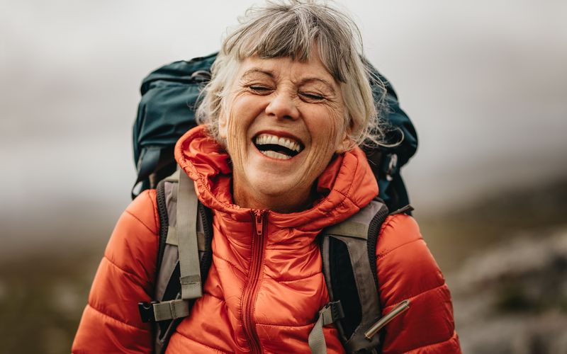 Close up of a senior woman wearing jacket and backpack laughing with eyes closed. Portrait of a woman hiker laughing during her trekking.